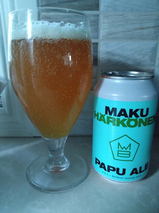 Ett glas med ljusbrun öl bredvid en ljusblå burk öl med texten "Maku Härkönen Papu Ale".