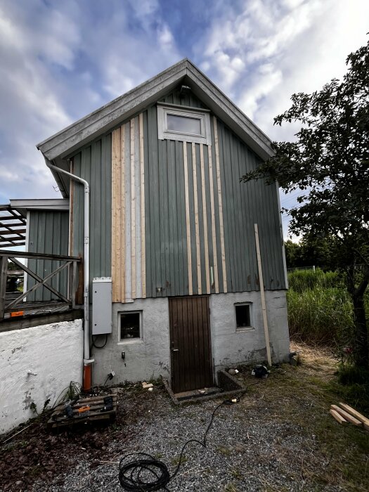 Sidorna av ett grönt uthyrningshus med ny träpanel och byggmaterial runt om. En gavel har börjat förberedas för målning med grundfärg.