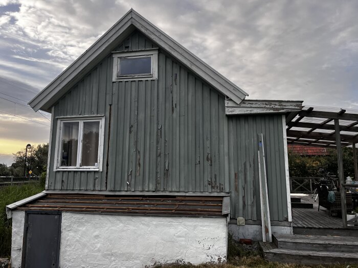 En sliten gröngrå trähusfasad med avflagnande färg, trasiga paneler och en nederdel med vitputsad tegel. En delvis färdig pergola syns till höger.