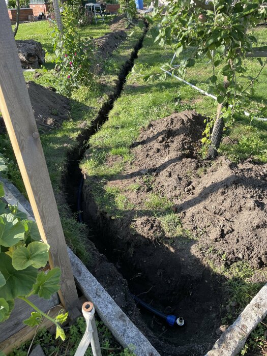 En grävd dike i en gräsmatta med nedlagd Pem-slang som kopplats till en vattenanslutning, omgiven av grönska och jordhögar.