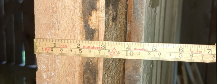 En mätsticka av trä med inches och centimeter används för att mäta tjockleken på en väggs träpanel, uppmätt till cirka 3 tum.