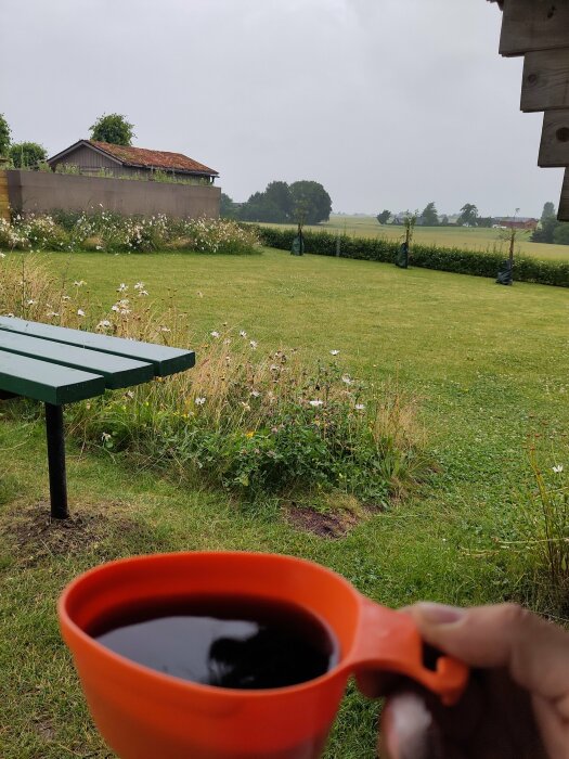 En person håller en orange kopp med kaffe framför en grön trädgård och en byggnad med rödmossigt tak i bakgrunden, under en mulen morgon med regn.