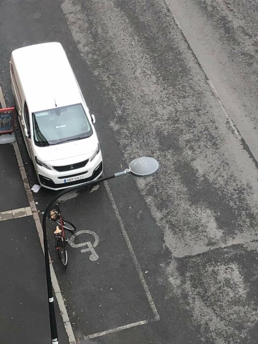 Vit skåpbil och cykel parkerade på en gata, skåpbilen står vid trottoarkanten och cykeln är placerad på en handikapparkering.