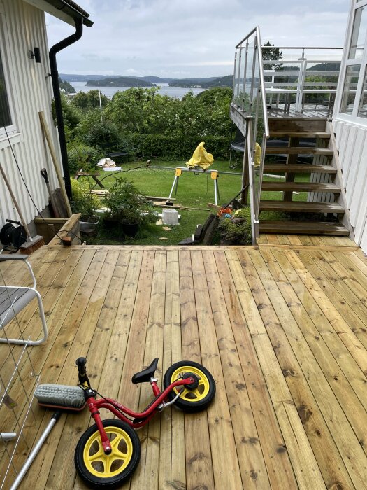 Nybyggd altan med en liten röd springcykel, en trappa som leder upp till en inglasad balkong och en trädgård med sjöutsikt i bakgrunden.