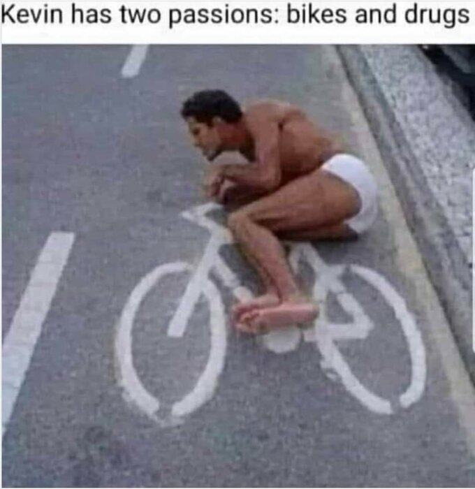 En man i underkläder ligger på marken och försöker imitera en cykel, med en vit cykelsymbol målad på asfalten under sig.