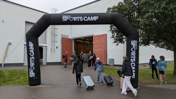 Deltagare anländer till Sportscamp, några bärande på bagage och andra med rullväskor, genom en stor uppblåsbar portal framför en byggnad med öppna portar.