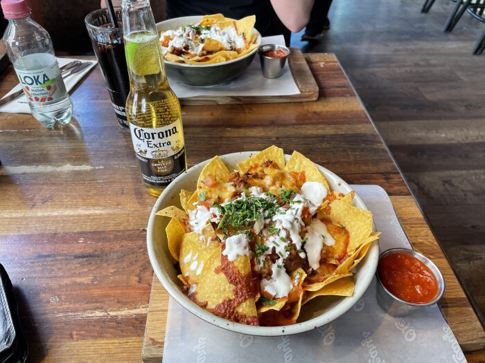 Skål med nachos toppade med gräddfil och örter, en liten skål med salsasås bredvid, Corona-öl samt vattenflaska och läsk i bakgrunden på ett träbord.
