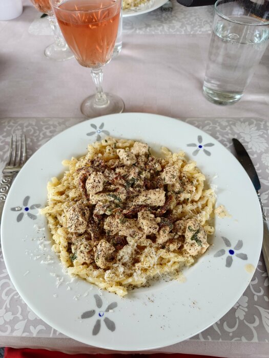 Tallrik med basilikakyckling och pasta toppad med parmesan och svartpeppar, bredvid ett glas rosévin och ett glas vatten på en dukad middag.