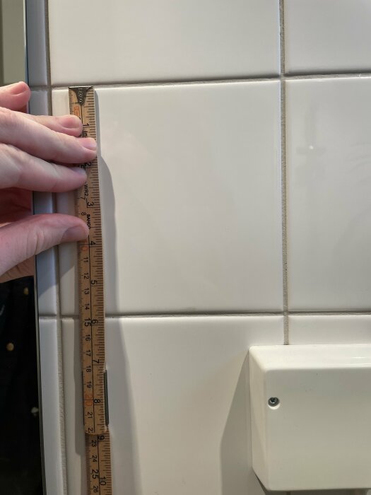 En linjal mäter storleken på vita kakelplattor i ett badrum, med en hand som håller linjalen bredvid kakelplattorna. En väggkontakt syns delvis i bilden.