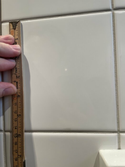 Vit kakelplatta mäts med tumstock i ett badrum, plattans höjd är ca 15 cm. En vit hand håller i tumstocken som är placerad parallellt med kakelplattans höjd.