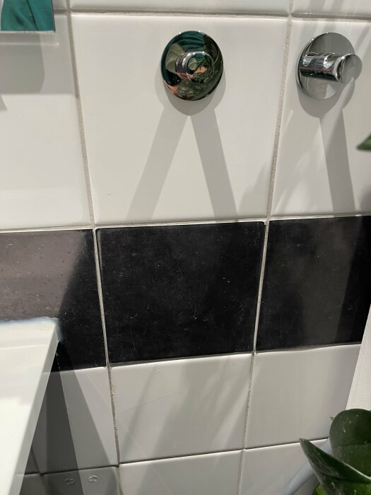Närbild på en vägg i ett badrum med svart och vitt kakel, två kromade krokar och en del av en vit bänkskiva synlig till vänster.