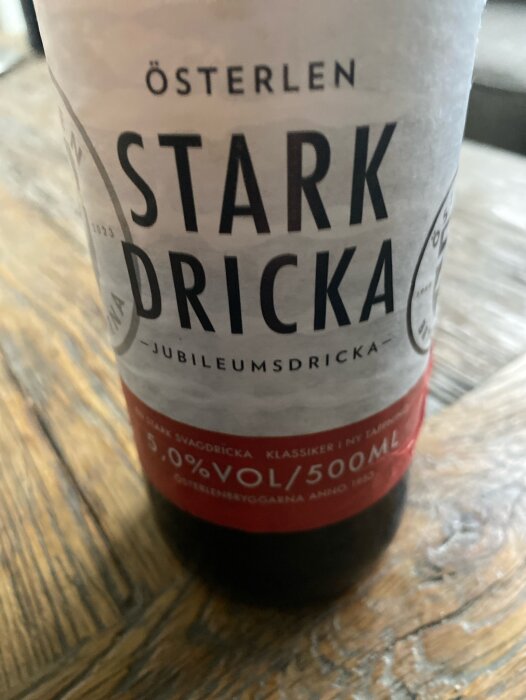 Flaska med etiketten "Österlen Starkdricka Jubileumsdricka" på ett träbord. Drickan har en alkoholhalt på 5.0% och en volym på 500 ml.