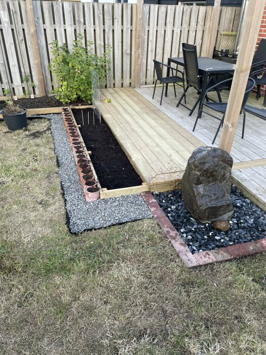 En trädgård med nyplanterade kryddor och tomatplantor, trätrall, stenar, möbler och en stor sten framför ett plank.