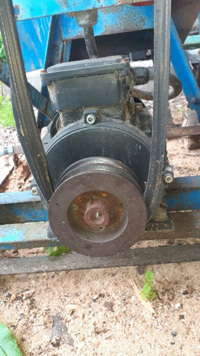 Närbild på svarta remmar och remskivor på en maskin med blå metallram i en verkstad, med träspån på marken.