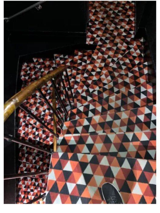 Trappa med geometriskt mönstrad matta i orange, svart och vitt sett ovanifrån. En fot med svart sko syns på första trappsteget.