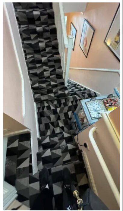 Svartvit trappa med geometriskt mönstrad matta, vita ledstänger och foton på väggarna. Skor och en trappskyffel är synliga i bilden.