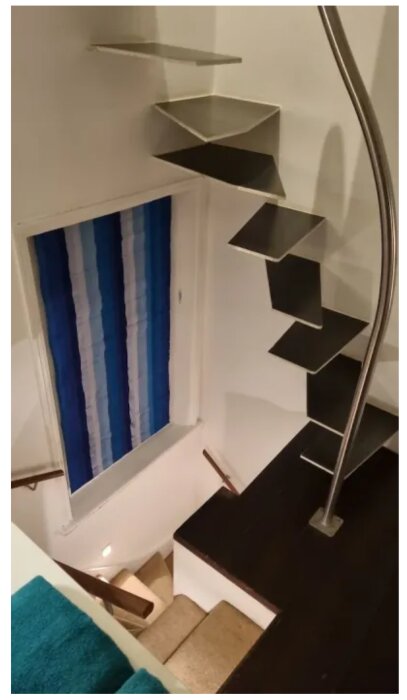 Modern svängd trappa i metall med flytande steg och räcke i rostfritt stål. Vägg med blå- och vitrandig gardin i bakgrunden.