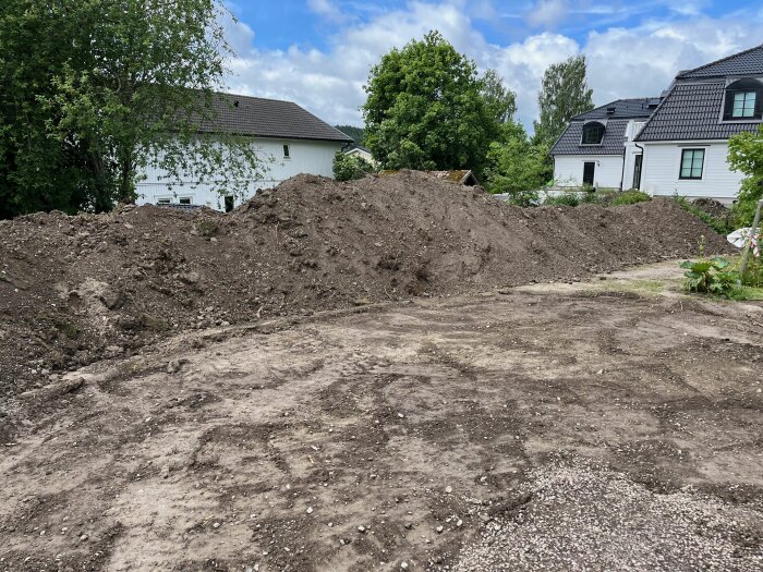 En stor hög med jord- och schaktmassor på en villatomt, redo för avhämtning. Hus och träd i bakgrunden.