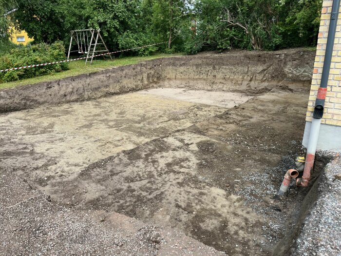 Utgrävd yta på en villatomt med uppgrävd matjord, lera och sand i Norrtälje, med ett rör och ett staket i bakgrunden.