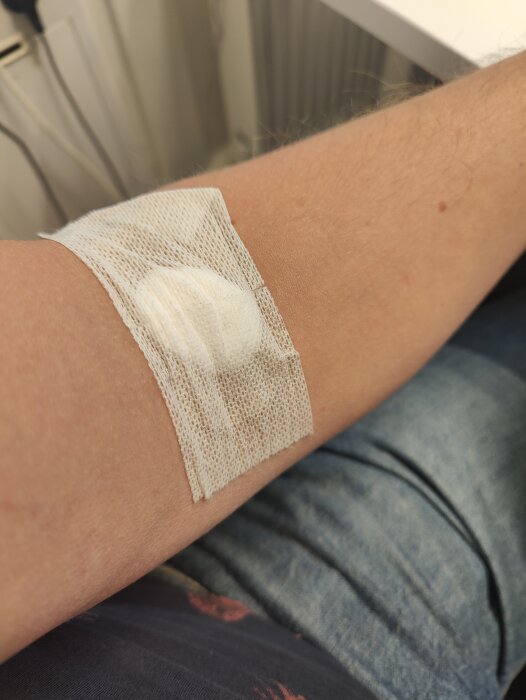 Armbåge med ett plåster och en gasbinda som täcker ett sår eller en injiceringspunkt på en persons arm.