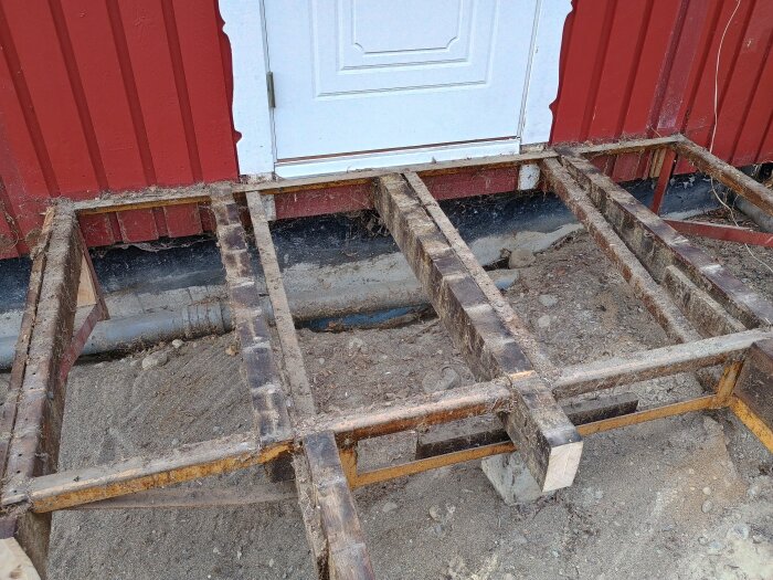 Ram av stål och trä för en rivd altan, monterad i backen framför en ytterdörr på ett rött hus. Detaljer av reglar och stöttor synliga.