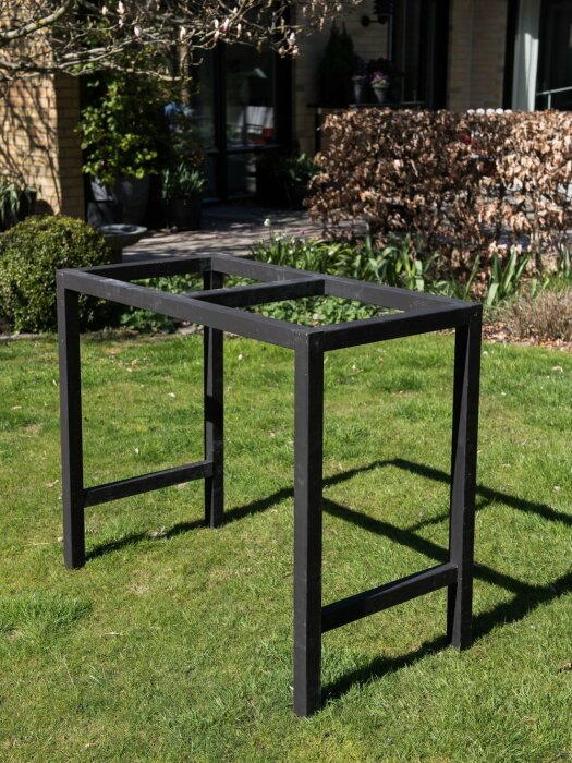 Svart sidobordsunderrede av 45x45 reglar stående på en gräsmatta i en trädgård. Underredet har inga synliga vinkeljärn eller urtag mellan reglarna.