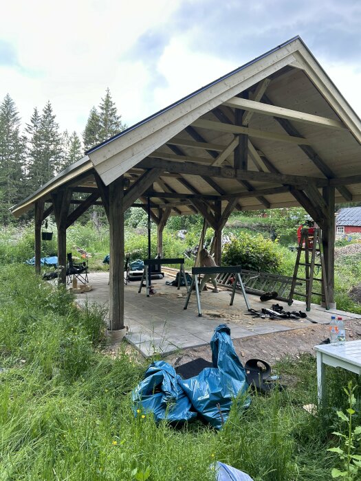 Grillpaviljong vid sommarhus byggd av återvunnet trä och ny råspont, omgiven av grönska och olika verktyg och material på marken.