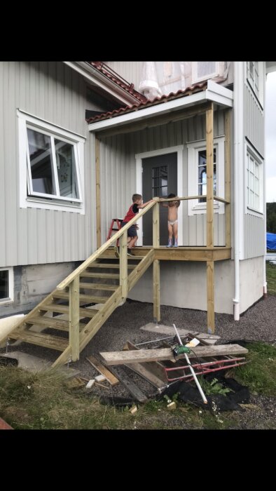 Två barn på en nybyggd trappa till en veranda vid ett grått hus. Byggmaterial ligger i en hög på marken bredvid trappan.