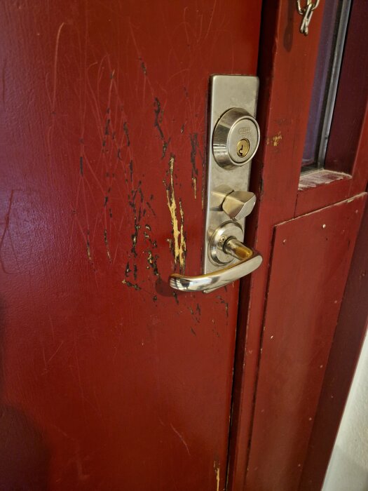 Röd trädörr med repor och skador runt ett silverfärgat dörrhandtag och lås. Skadan är mest koncentrerad precis vid dörrhandtaget.