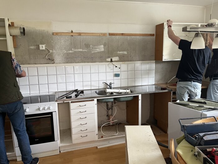 Renovering av ett kök där två personer arbetar, en med att montera köksskåp och den andra med att fixa ovanskåp. Väggen är delvis utan kakel och verktyg ligger på bänken.