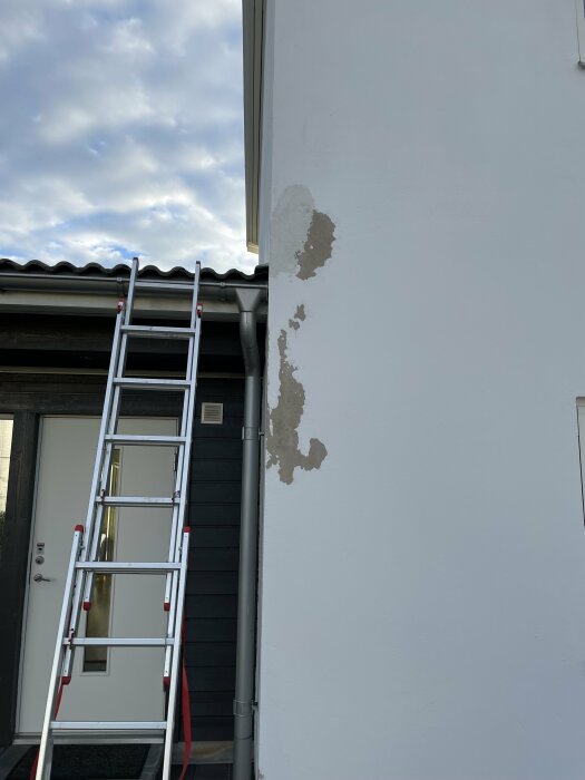 Färgen lossnar på husets fasad vid ett hörn, en stege står lutad mot väggen bredvid dörren. Molning himmel i bakgrunden.