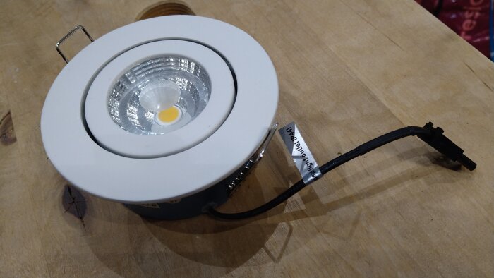 LED-spotlight med vit ram, placerad på en träyta med en svart kabel lödad direkt på LED-enheten och en kontaktanslutning.