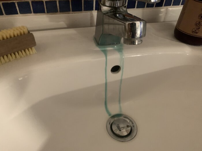 Tvättställ med vattenkran som har en grönaktig beläggning som rinner från fogen. En tvättborste och en flaska är placerade bredvid.