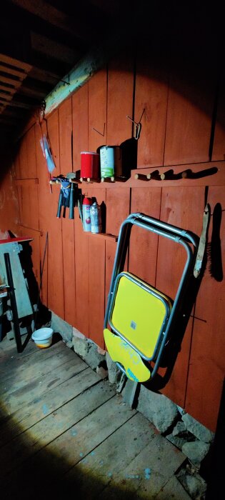 Verkstad med olika verktyg och färgburkar som hänger på krokar på en röd trävägg. Ett gult hopfällbart bord står lutat mot väggen.