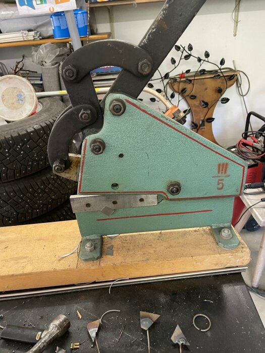 Grön industrimaskin med handtag, monterad på en träbas, omgiven av verktyg och däck i ett garage.