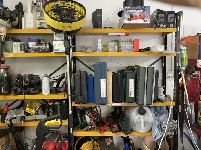 Hyllor med olika verktyg och maskiner, inklusive borrmaskiner, förvaringslådor, burkar med kemikalier och andra byggtillbehör i ett garage.