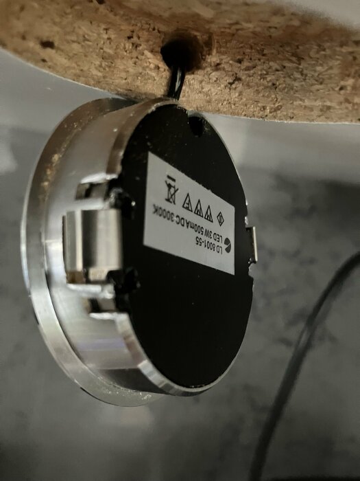 LED-puck fäst under ett skåp med en svart kabel som går igenom ett hål. Pucken har en etikett med produktinformation och varningssymboler på baksidan.