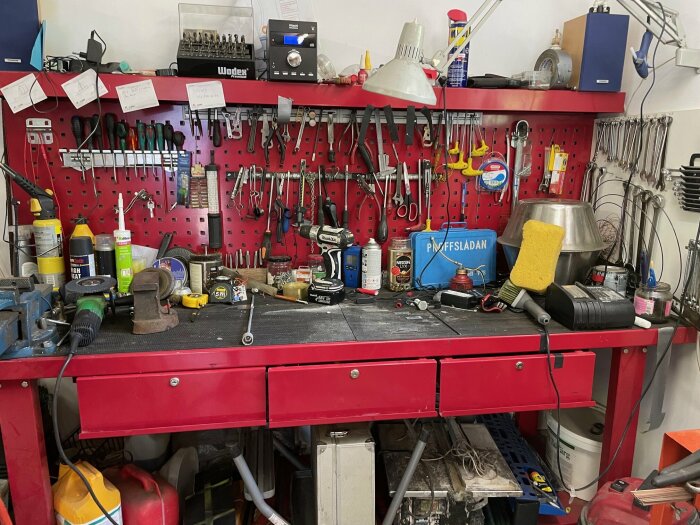 Rött arbetsbord med en röd verktygstavla på väggen i ett garage, fullt med olika verktyg och redskap, inklusive borrmaskin, skruvmejslar och andra handverktyg.