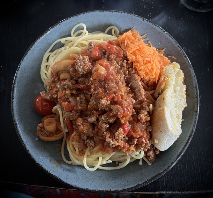 Tallrik med spaghetti och köttfärssås, serverad med rivna morötter och en skiva vitlöksbröd.