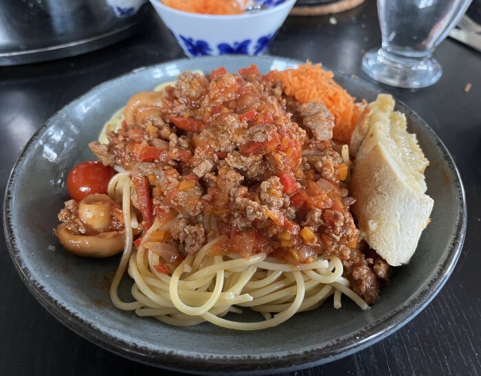 Tallrik med spaghetti och köttfärssås, serverad med rivna morötter och vitlöksbröd; glas och en skål med morötter i bakgrunden.
