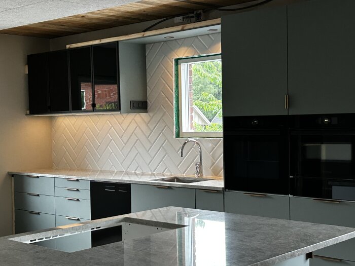 Modernt kök med grå bänk, mönstrat kakel, inbyggd belysning, och fönster ovanför diskbänken. Knappar syns till vänster om fönstret.