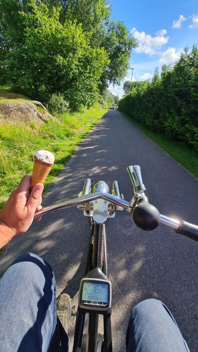 Person cyklar på en landsväg medan de håller en glass i ena handen och cykelstyret i den andra, omgiven av grönska och ett klart himmel.