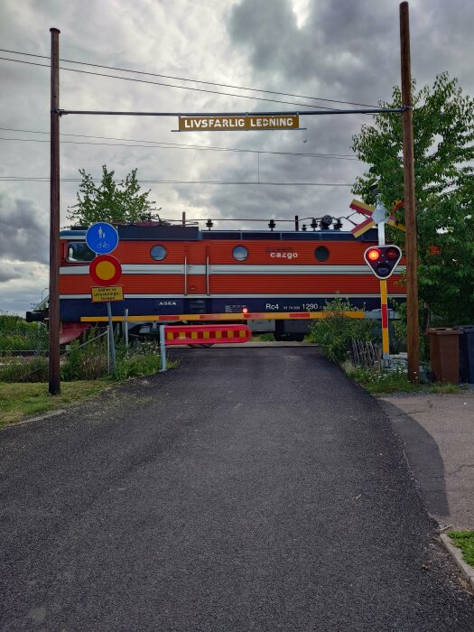 Ett orange och blått godståg går över en järnvägskorsning med en skylt som varnar för livsfarlig ledning ovanför, tillsammans med ett rött stoppljus och cykelskylt.