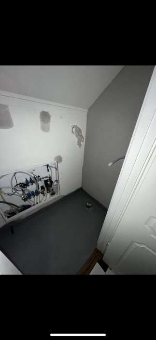 Skåp för golvvärmeslingor installerat på väggen i en liten toalett, med rör och komponenter synliga. Väggen är delvis vit och delvis gråmålad.