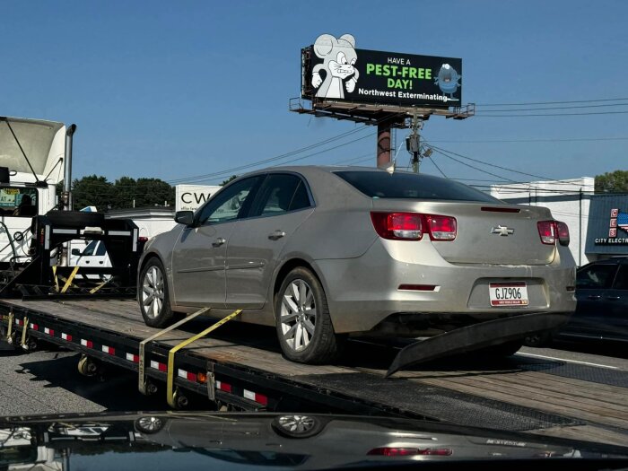 Bil med skador på bakre stötfångare lastad på en trailer, i bakgrunden syns en reklamskylt med texten "Have a pest-free day! Northwest exterminating".