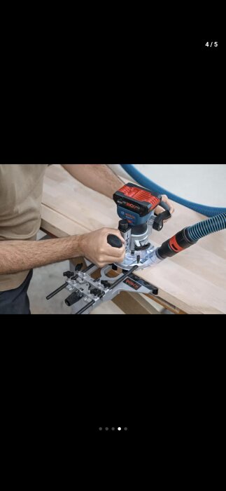 En person använder en Bosch 18V handöverfräs för träbearbetning på en arbetsbänk, med en dammsugarslang ansluten.
