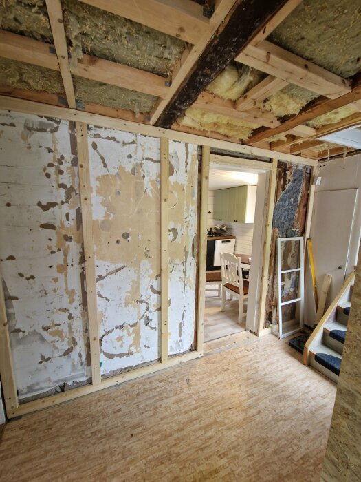 En hall under renovering med riven masonit och uppsatt gles på väggen, samt gammal isolering synlig i taket. En öppen dörr leder till köket.