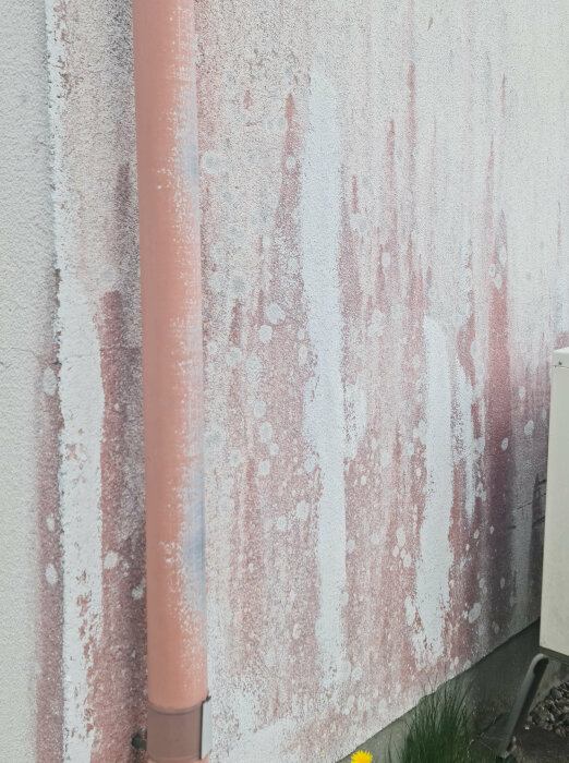 Putsad fasad med färgskiftningar och restspår av borttagna rödalger. En beige rörledning löper vertikalt längs väggen till vänster i bilden.