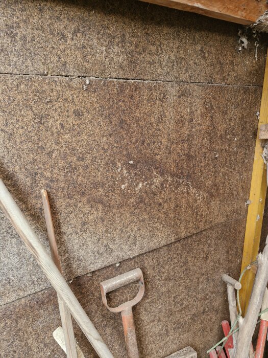 Vägg av bruna skivor på en altan, med några verktyg som står lutade mot väggen, inklusive en spade.