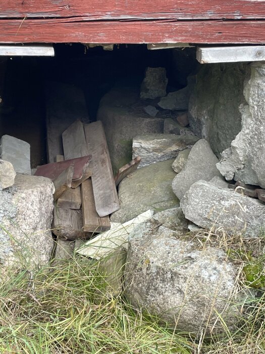 Husets hörn med borttagna stenar, synliga träbjälkar och grus under. Grass och stenar syns i förgrunden.